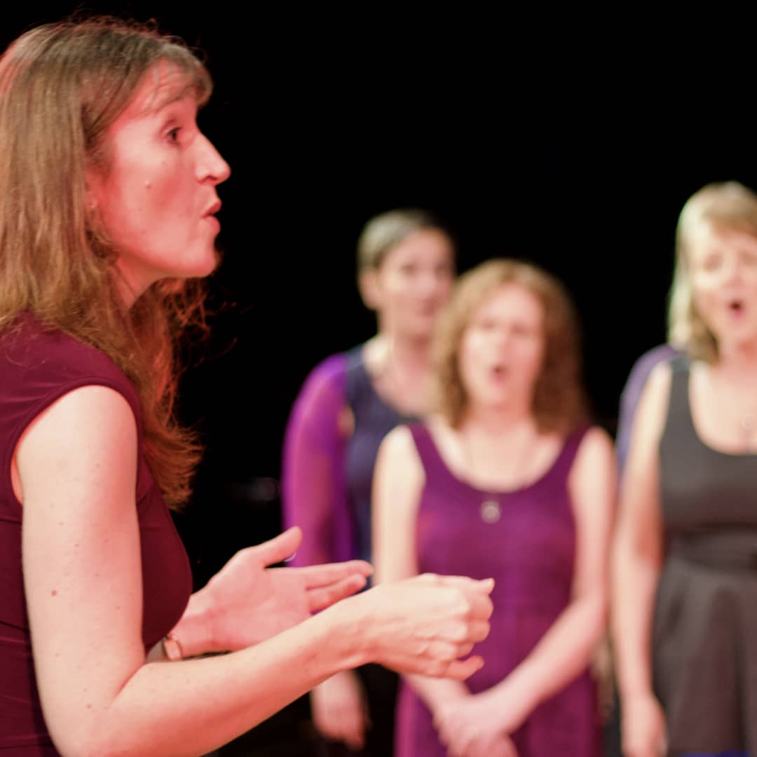 Close-up of Imogen Wolf conducting WolfSong choir at Byron Bay Theatre, singers in background performing ‘oooh’ sound