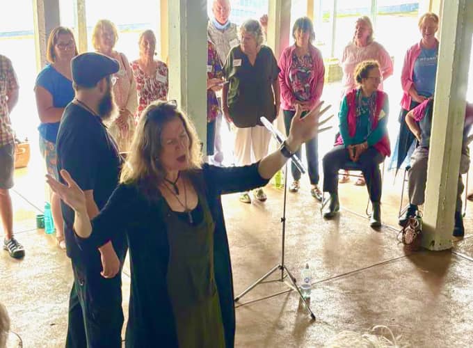 Imogen Wolf singing with arms raised while Brian Martin and a group of participants stand in a semicircle beneath the Maleny Community Centre