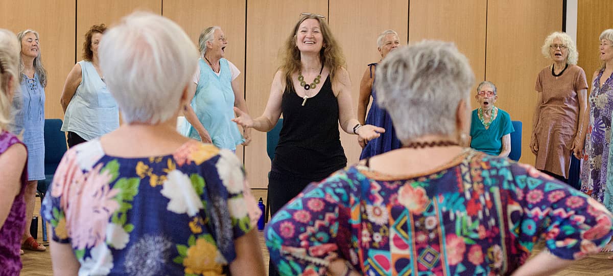 Imogen Wolf standing at the centre of a singing circle indoors, smiling and gesturing as participants sing around her