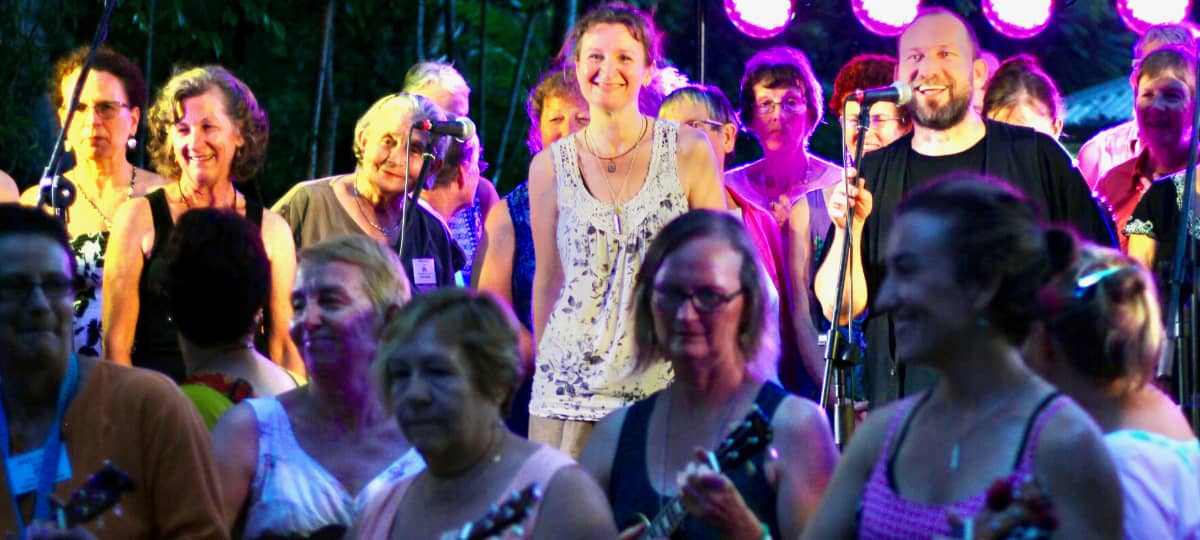 Camp Creative Party In The Park concert with ukulele players in the foreground and Joy Of Singing choir at the back under stage lights"