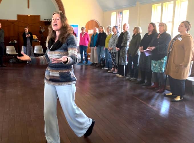 Imogen Wolf guiding a group of adult singers during a vocal workshop in a sunlit community hall.