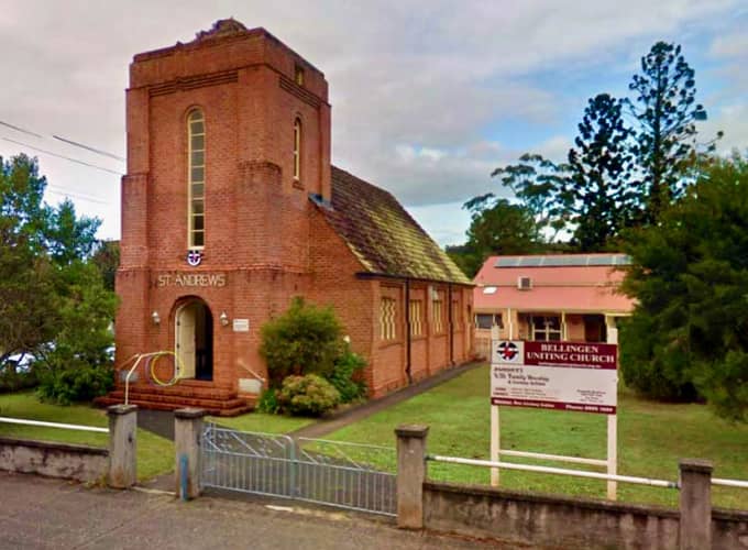 Brick church building with tower and sign reading “Bellingen Uniting Church,” surrounded by greenery.