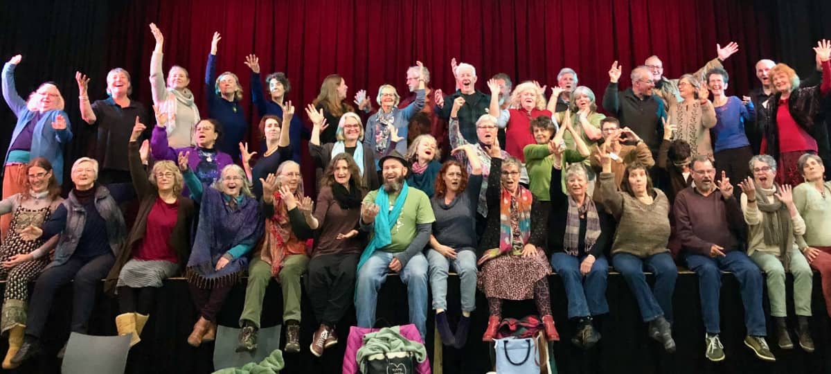 Large group of WolfSong winter camp participants seated and standing on a theatre stage with red curtain backdrop, arms raised